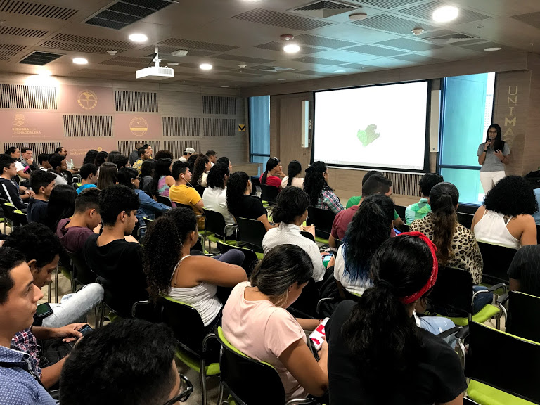 A group of at least 40 students are sitting in a lecture room listening to a presentation. A large screen in the room projects the slides for the presentation. © M. Smith.