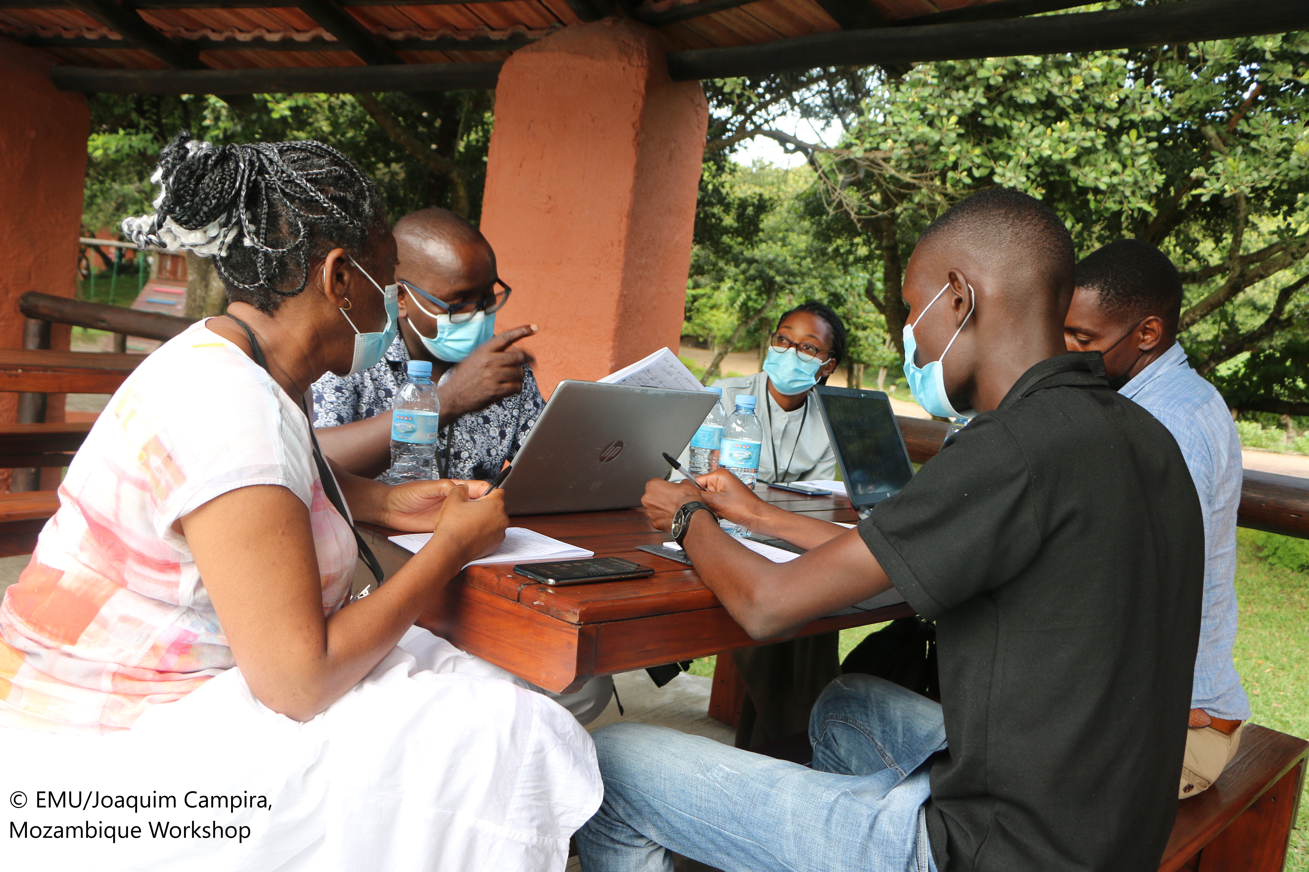 Photograph of a group of five people sitting around an outdoor table with laptops and notepads 
