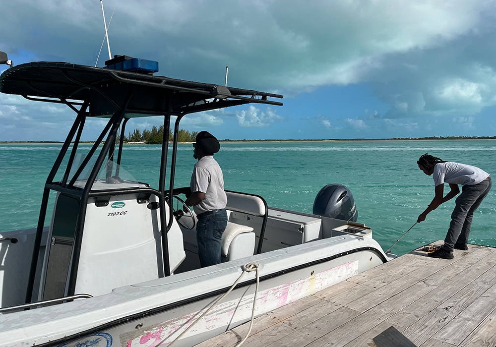 Photograph of a small boat leaving a dock, with one man driving and another pushing it away (copyright Jane Hawkridge)