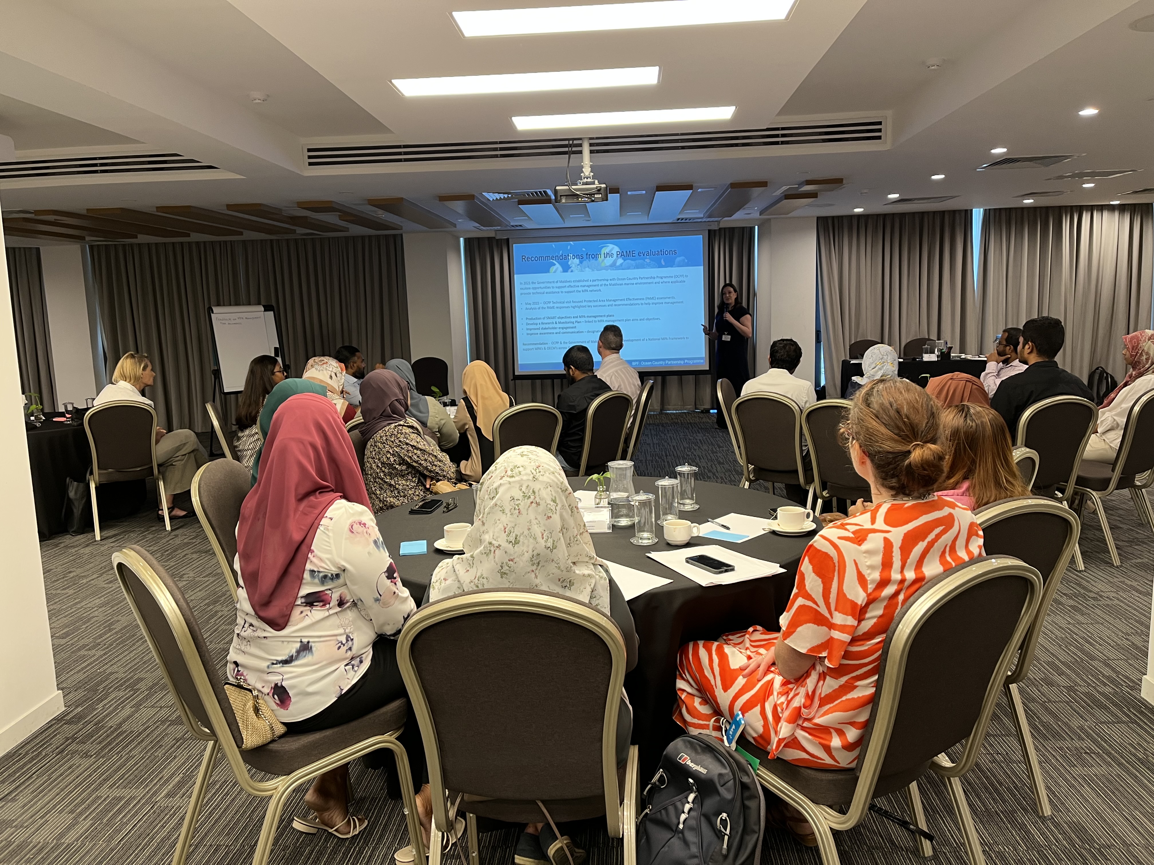 A room of stakeholders sitting at round tables watching a presentation for a workshop.