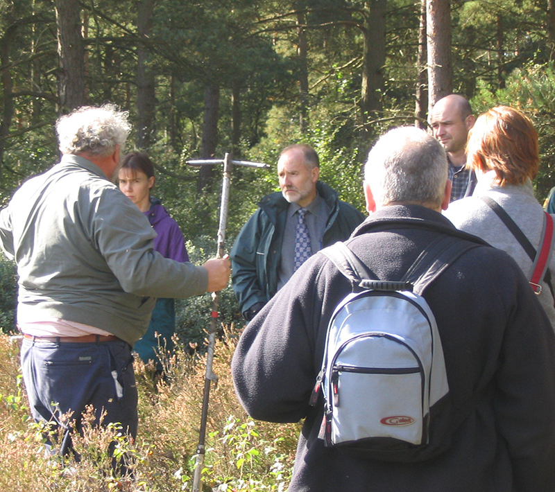 Photograph of a group of 6 volunteers carrying out some monitoring. The group are in scrubland close to some trees. One of them is holding some monitoring equipment. 
