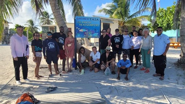 Photo of group standing in front of sign on a beach (OCPP, IUCN and Belize MPA managers and comanagers on a field visit to TASA headquarters, Turneffe Atoll)