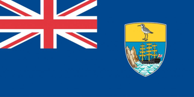 The Flag of St Helena. The flag is a deep shade of blue. In the top left features the UK flag with blue, red and white stripes. On the right hand side is a shield split into two. In the top half, a St Helena Plover sits on a yellow background. The bottom half is an old style ship sailing up to some cliffs