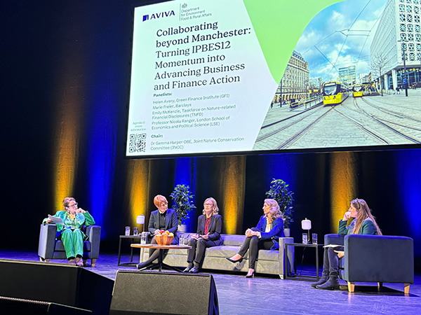 Photograph of the Aviva/Defra Business & Finance Day, showing a close-up of a group of 5 panel members seated in a row and in discussion. Behind and above them is a large screen displaying a presentation. 